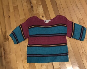 May include: A short-sleeved sweater with horizontal stripes in teal, burgundy, and black. The sweater has a round neckline and a ribbed hem and cuffs. The color scheme is retro, with a vintage aesthetic.