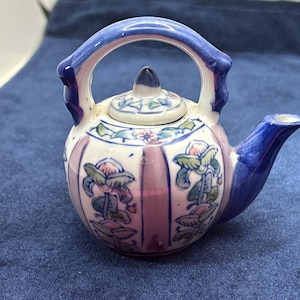 May include: A small ceramic teapot featuring a rounded body with vertical pink stripes and hand-painted green and pink floral motifs. The teapot has a blue handle, a matching blue spout, and a removable lid with a pointed finial top.