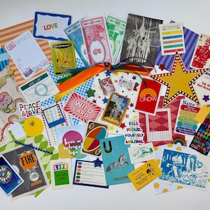 May include: A collection of colorful paper ephemera, game cards, stickers, and printed notes featuring various illustrations, text, and patterns. The items are arranged in a scattered cluster and tied together with a multi-colored ribbon.
