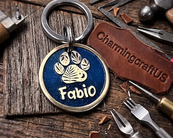 May include: A round metal pet tag with a dark blue resin center featuring a gold-colored paw print design with leaf patterns inside the paw pads. The name Fabio is printed in gold letters below the paw print. The tag has a silver metal keyring.