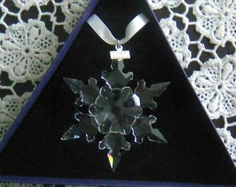 May include: A clear crystal snowflake ornament with a silver ribbon, presented in a triangular black box. The snowflake has six points and a small silver tag with the year 2022. The background is a white lace pattern.