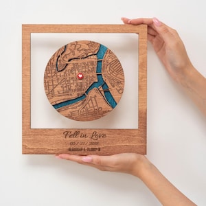 Puede incluir: Una obra de arte de mapa de madera con un dise&ntilde;o de mapa de ciudad circular, con un marcador de coraz&oacute;n rojo. El mapa est&aacute; sobre un fondo azul, enmarcado en un marco de madera marr&oacute;n claro. El texto "Fell in Love" y una fecha est&aacute;n grabados debajo.