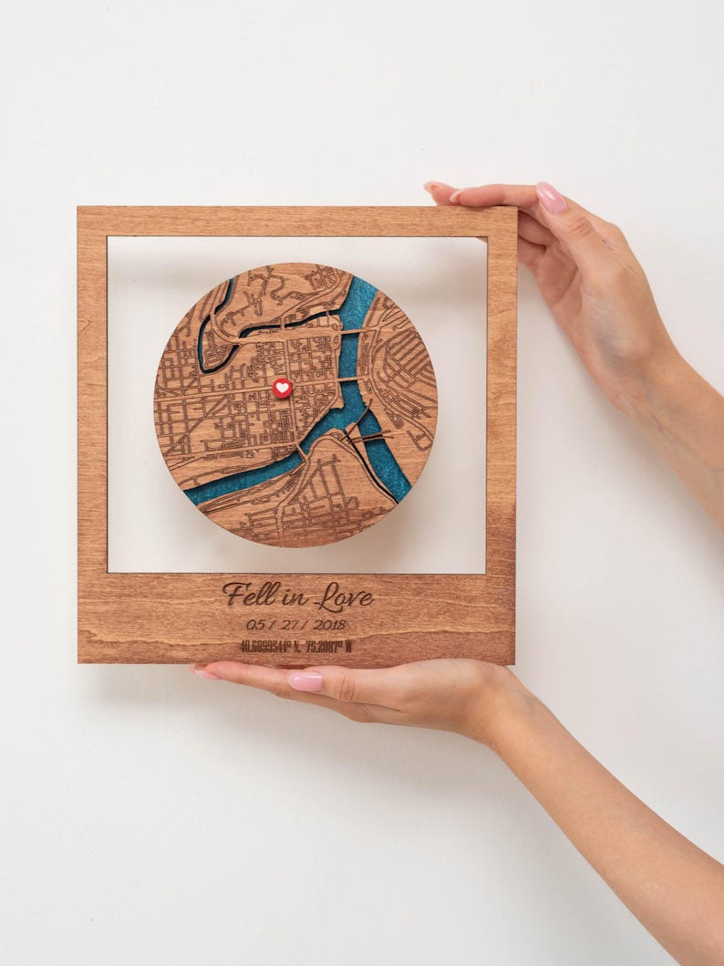 Puede incluir: Una obra de arte de mapa de madera con un dise&ntilde;o de mapa de ciudad circular, con un marcador de coraz&oacute;n rojo. El mapa est&aacute; sobre un fondo azul, enmarcado en un marco de madera marr&oacute;n claro. El texto "Fell in Love" y una fecha est&aacute;n grabados debajo.