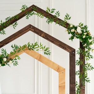 May include: A wooden archway decorated with greenery and white flowers. The archway is made of two pieces of wood, one brown and one natural wood.
