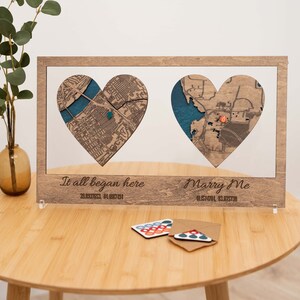 Puede incluir: Obra de arte enmarcada en madera con dos mapas en forma de coraz&oacute;n y detalles azules. El marco incluye el texto "It all began here" y "Marry Me" con coordenadas. Una mesa peque&ntilde;a sostiene la obra y tarjetas decorativas.