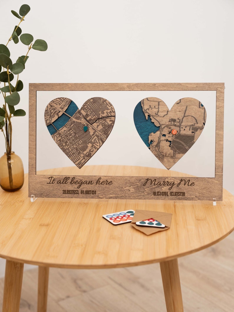 Puede incluir: Obra de arte enmarcada en madera con dos mapas en forma de coraz&oacute;n y detalles azules. El marco incluye el texto "It all began here" y "Marry Me" con coordenadas. Una mesa peque&ntilde;a sostiene la obra y tarjetas decorativas.