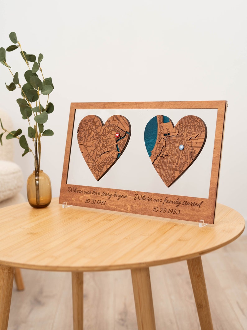Puede incluir: Obra de arte enmarcada en madera con dos recortes de mapas en forma de coraz&oacute;n. Un coraz&oacute;n tiene un alfiler rojo, el otro un alfiler azul. El texto dice "Where our love story began 10.31.1981" y "Where our family started 10.29.1983".