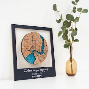 Puede incluir: Un mapa de madera enmarcado con un dise&ntilde;o circular, que presenta un plano detallado de la ciudad con un r&iacute;o azul y un peque&ntilde;o marcador de color cobre. El marco es azul oscuro, y el texto "Where we got engaged" se muestra debajo, junto con la fecha y las coordenadas.