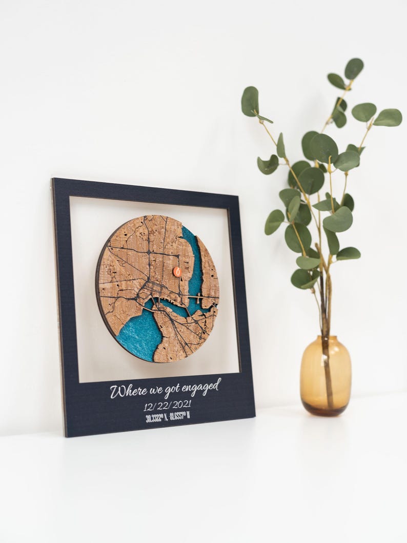 Puede incluir: Un mapa de madera enmarcado con un dise&ntilde;o circular, que presenta un plano detallado de la ciudad con un r&iacute;o azul y un peque&ntilde;o marcador de color cobre. El marco es azul oscuro, y el texto "Where we got engaged" se muestra debajo, junto con la fecha y las coordenadas.