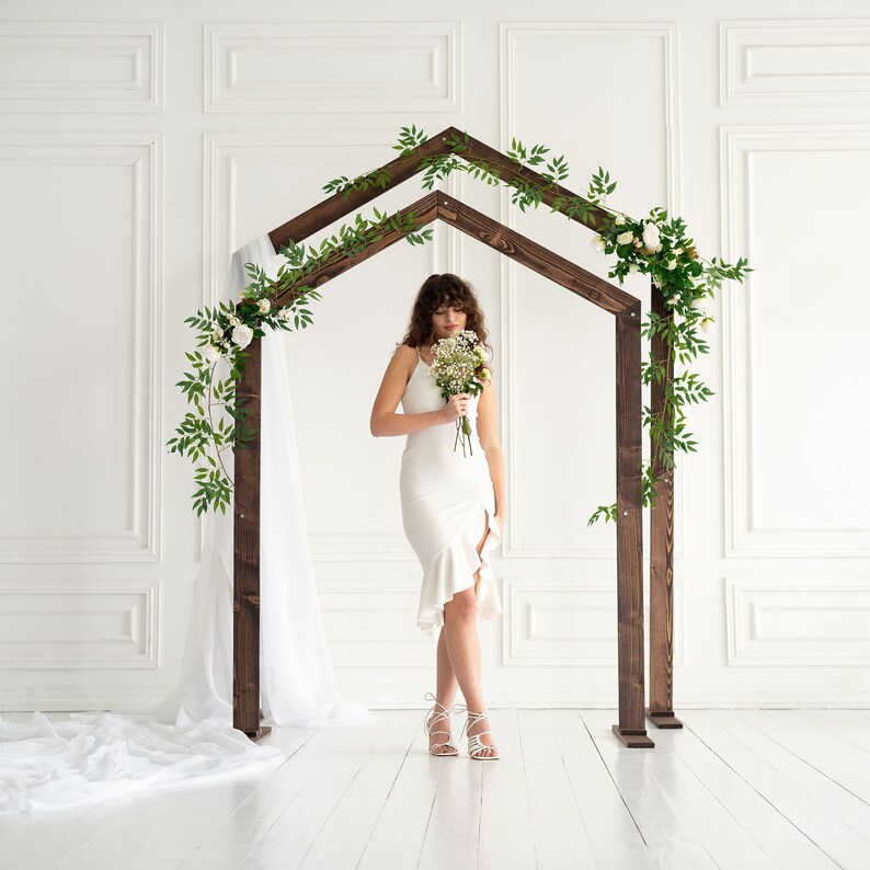 May include: A white wedding dress with a ruffled hem and a bouquet of white flowers. The bride is standing under a wooden archway decorated with greenery and white flowers.