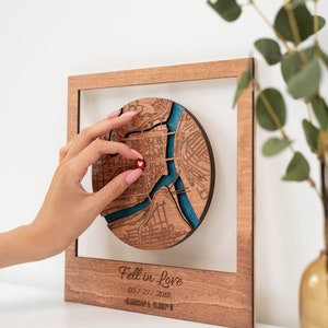 Puede incluir: Arte de mapa de madera con un dise&ntilde;o circular de mapa de la ciudad, con un r&iacute;o azul y un coraz&oacute;n rojo. El mapa est&aacute; dentro de un marco de madera, con el texto "Fell in Love" y una fecha. Una pieza de decoraci&oacute;n del hogar &uacute;nica.