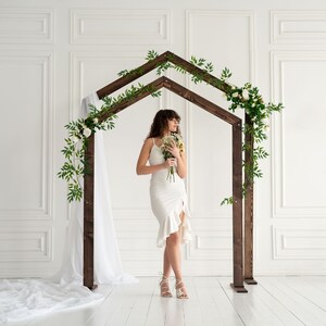 May include: A white wedding dress with a ruffled hemline, worn by a person standing under a wooden archway decorated with greenery and white flowers.