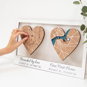 Puede incluir: Una obra de arte enmarcada con dos mapas de madera en forma de coraz&oacute;n. Un coraz&oacute;n tiene un alfiler rojo, el otro un acento azul. El texto "Bounded by love" y "Our First Home" est&aacute;n debajo, junto con coordenadas.