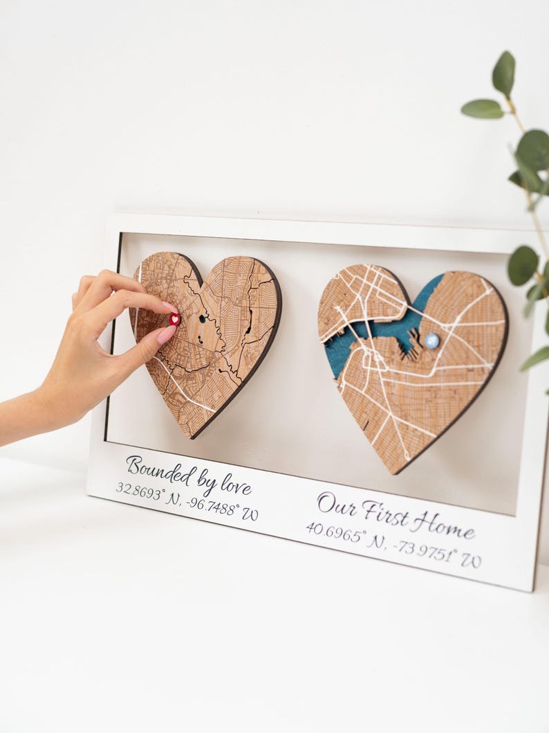 Puede incluir: Una obra de arte enmarcada con dos mapas de madera en forma de coraz&oacute;n. Un coraz&oacute;n tiene un alfiler rojo, el otro un acento azul. El texto "Bounded by love" y "Our First Home" est&aacute;n debajo, junto con coordenadas.
