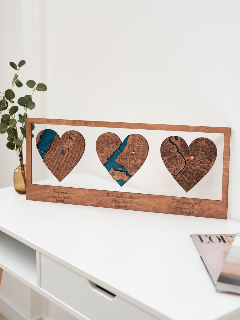 Puede incluir: Obra de arte de madera con tres mapas en forma de coraz&oacute;n. Cada coraz&oacute;n presenta un mapa de la ciudad con un elemento de agua azul turquesa. El texto debajo de cada coraz&oacute;n dice "We met December 2022 New York", "We fell in love May 2023 Niagara Falls" y "We engaged September 2023 Philadelphia".