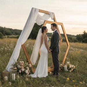 May include: A large wooden hexagonal wedding arch decorated with white fabric drapes. A person in a white floor-length gown with a veil and a person in a dark gray suit stand facing each other under the arch, surrounded by floral arrangements and candles.