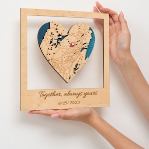Puede incluir: Un mapa de madera en forma de coraz&oacute;n con un alfiler rojo, enmarcado en un marco de madera clara. El mapa est&aacute; detallado con l&iacute;neas grabadas y se asienta sobre un fondo turquesa. El marco incluye el texto grabado "Together, always yours" y la fecha 6/15/2023.