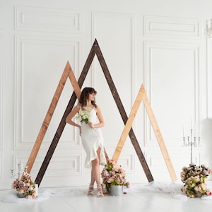 Può includere: Un set di tre grandi cornici triangolari in legno in varie tonalità di marrone, disposte in una formazione sovrapposta simile a una montagna. La struttura è decorata con bouquet di fiori rosa e bianchi e candelabri argentati sul pavimento.