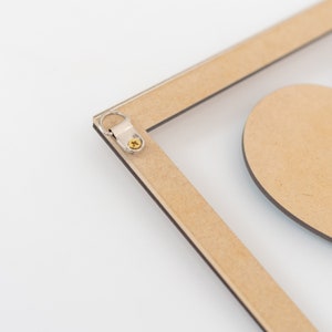 Puede incluir: Primer plano de un marco de fotos de madera con una inserci&oacute;n de acr&iacute;lico transparente. El marco tiene un colgador de metal sujeto con un tornillo. Una forma de coraz&oacute;n de madera es visible en el fondo. El marco es de color marr&oacute;n claro y el fondo es blanco.