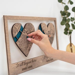 Puede incluir: Obra de arte enmarcada en madera con dos recortes de mapas en forma de coraz&oacute;n y respaldo color turquesa. La frase "It all began here" est&aacute; grabada en la parte inferior, junto con las coordenadas. Una mano interact&uacute;a con la obra de arte. La obra se exhibe en una pared blanca.