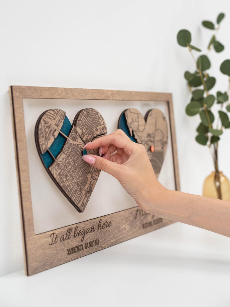 Puede incluir: Obra de arte enmarcada en madera con dos recortes de mapas en forma de coraz&oacute;n y respaldo color turquesa. La frase "It all began here" est&aacute; grabada en la parte inferior, junto con las coordenadas. Una mano interact&uacute;a con la obra de arte. La obra se exhibe en una pared blanca.