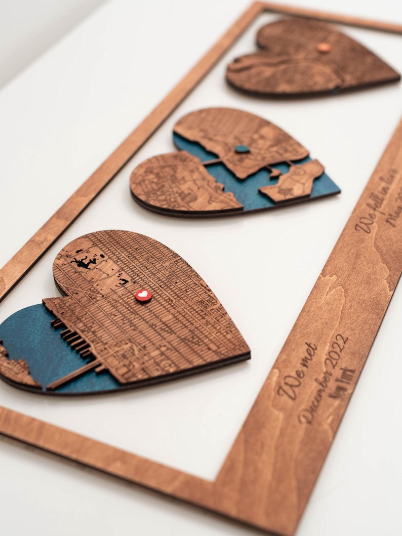 Puede incluir: Arte de mapa en forma de coraz&oacute;n de madera con un marco. El arte presenta tres mapas en forma de coraz&oacute;n con un fondo azul. El marco tiene el texto "We met December 2022 New York" y "We fell in love May 2023".