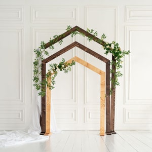 May include: A wooden archway decorated with greenery and white flowers. The archway is made of two different types of wood, one dark brown and one light brown. The archway is standing in front of a white wall with decorative molding.