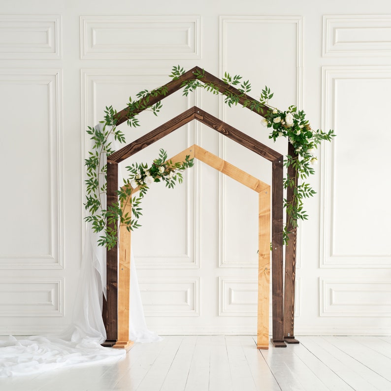 May include: A wooden archway decorated with greenery and white flowers. The archway is made of two different types of wood, one dark brown and one light brown. The archway is standing in front of a white wall with decorative molding.