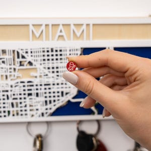 May include: A small circular red pin featuring a white icon of a hand holding a wine glass. In the background, a white wooden wall-mounted key holder displays a map of Miami with the word MIAMI in large white letters above the map grid.