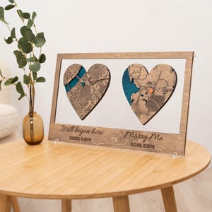 Puede incluir: Obra de arte enmarcada en madera con dos recortes de mapas en forma de coraz&oacute;n y detalles turquesa. Los textos "It all began here" y "Marry Me" est&aacute;n grabados debajo de los mapas, junto con las coordenadas. La obra se exhibe sobre una mesa de madera.