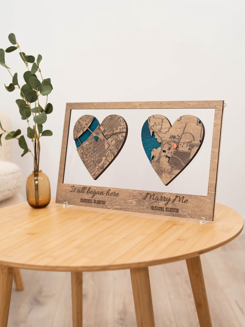 Puede incluir: Obra de arte enmarcada en madera con dos recortes de mapas en forma de coraz&oacute;n y detalles turquesa. Los textos "It all began here" y "Marry Me" est&aacute;n grabados debajo de los mapas, junto con las coordenadas. La obra se exhibe sobre una mesa de madera.