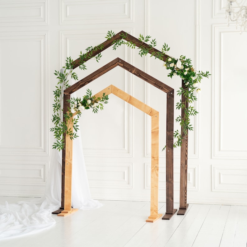 May include: A wooden wedding arch with two tiers, the top tier is dark brown and the bottom tier is light brown. The arch is decorated with greenery and white flowers.