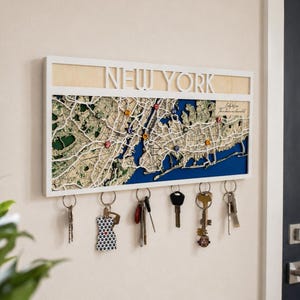 May include: A rectangular wall-mounted wooden key holder featuring a layered map of New York with the text NEW YORK at the top. The map includes blue water sections, green land areas, and small colored markers. Six metal hooks hold various keys.