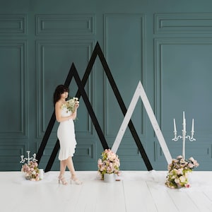 May include: A bride in a white dress stands in front of a teal wall with black and white geometric shapes. She is holding a bouquet of white flowers. There are two floral arrangements on either side of her.