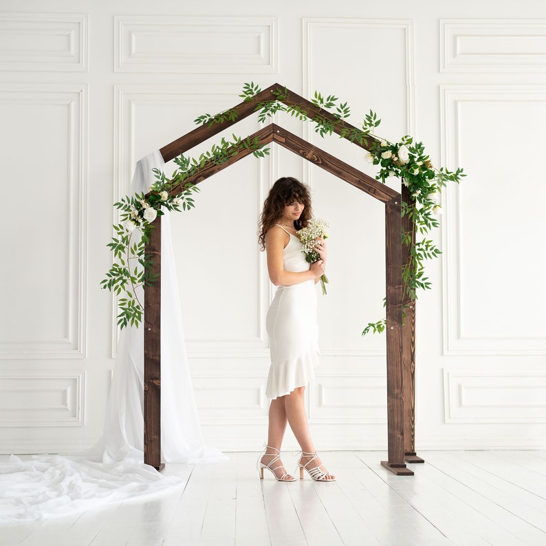 May include: A woman in a white dress stands under a wooden archway decorated with greenery and white flowers. The archway is in a white room with white walls and a white floor.
