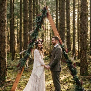 May include: A triangular wooden wedding arch decorated with pine branches, pinecones, and red berries. A person in a white lace gown with bell sleeves and a floral crown stands facing a person in a brown tweed suit, holding hands in a forest.