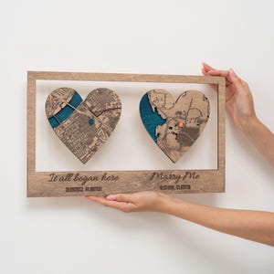 Puede incluir: Una obra de arte enmarcada en madera con dos mapas en forma de coraz&oacute;n y detalles turquesa. Las frases "It all began here" y "Marry Me" est&aacute;n grabadas debajo. La obra se presenta sobre un fondo blanco.