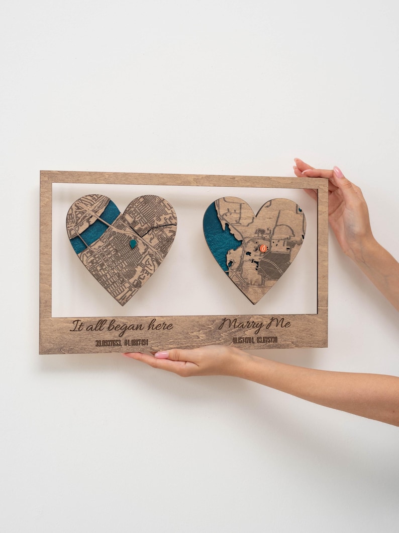 Puede incluir: Una obra de arte enmarcada en madera con dos mapas en forma de coraz&oacute;n y detalles turquesa. Las frases "It all began here" y "Marry Me" est&aacute;n grabadas debajo. La obra se presenta sobre un fondo blanco.