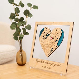 Puede incluir: Un mapa en forma de coraz&oacute;n de madera con un alfiler rojo, enmarcado en un marco de madera clara. El mapa tiene un fondo azul y calles detalladas. El marco incluye el texto "Together, always yours" y la fecha 15/06/2023.