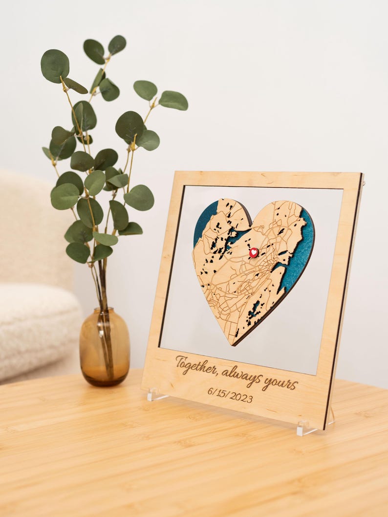 Puede incluir: Un mapa en forma de coraz&oacute;n de madera con un alfiler rojo, enmarcado en un marco de madera clara. El mapa tiene un fondo azul y calles detalladas. El marco incluye el texto "Together, always yours" y la fecha 15/06/2023.