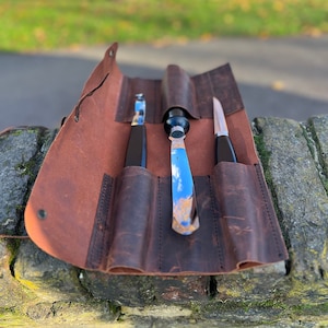 May include: A brown leather roll with three slots for holding tools. The roll is open and shows three tools, two with black handles and one with a silver handle.