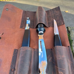May include: A brown leather roll with three slots for holding tools. The roll is closed and the tools are visible inside. The tools are a small curved knife, a larger curved knife, and a straight knife. All three knives have black handles.