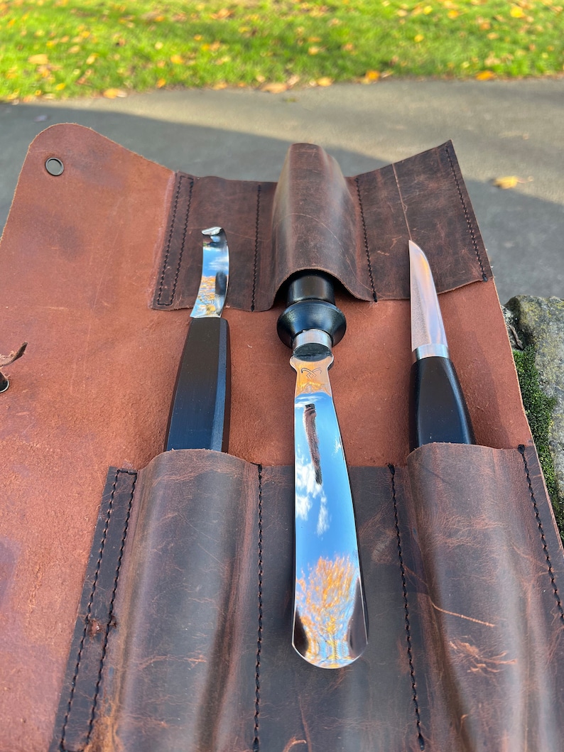 May include: A brown leather roll with three slots for holding tools. The roll is closed and the tools are visible inside. The tools are a small curved knife, a larger curved knife, and a straight knife. All three knives have black handles.
