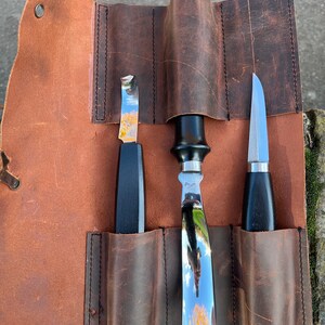 May include: A brown leather roll with three slots for tools. The roll is open and shows three tools: a small, curved blade, a larger, straight blade, and a small, pointed blade. All three tools have black handles.