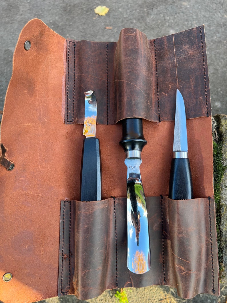 May include: A brown leather roll with three slots for tools. The roll is open and shows three tools: a small, curved blade, a larger, straight blade, and a small, pointed blade. All three tools have black handles.