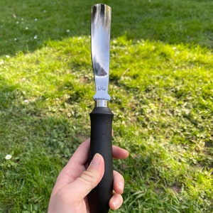 May include: A black handled stainless steel scraper tool with a curved blade. The tool is held in a hand against a green grassy background.