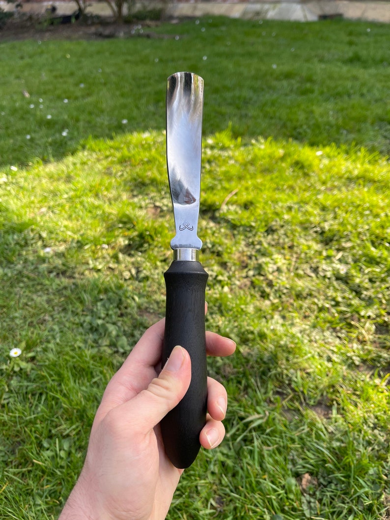 May include: A black handled stainless steel scraper tool with a curved blade. The tool is held in a hand against a green grassy background.