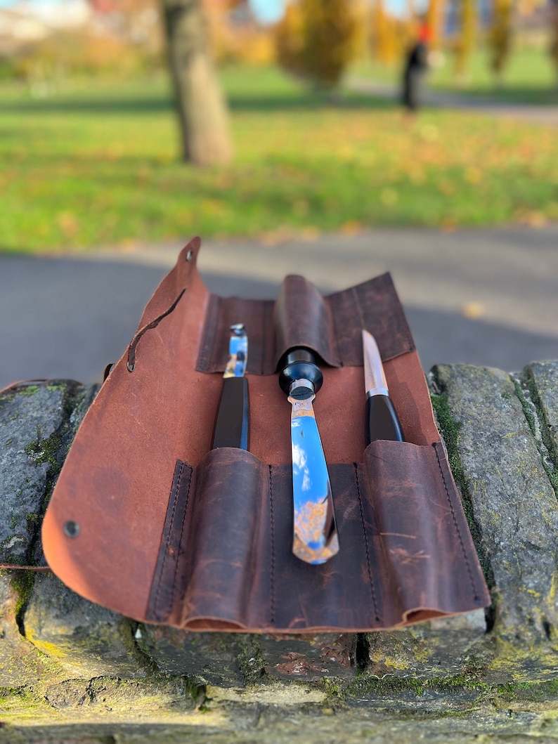 May include: A brown leather roll with three slots for holding tools. The roll is open and shows three tools, two with black handles and one with a silver handle.
