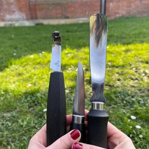 May include: Three different knives with black handles. The knives have silver blades and are held in a person's hand.