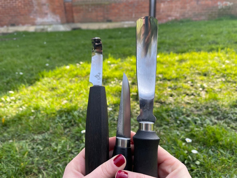May include: Three different knives with black handles. The knives have silver blades and are held in a person's hand.